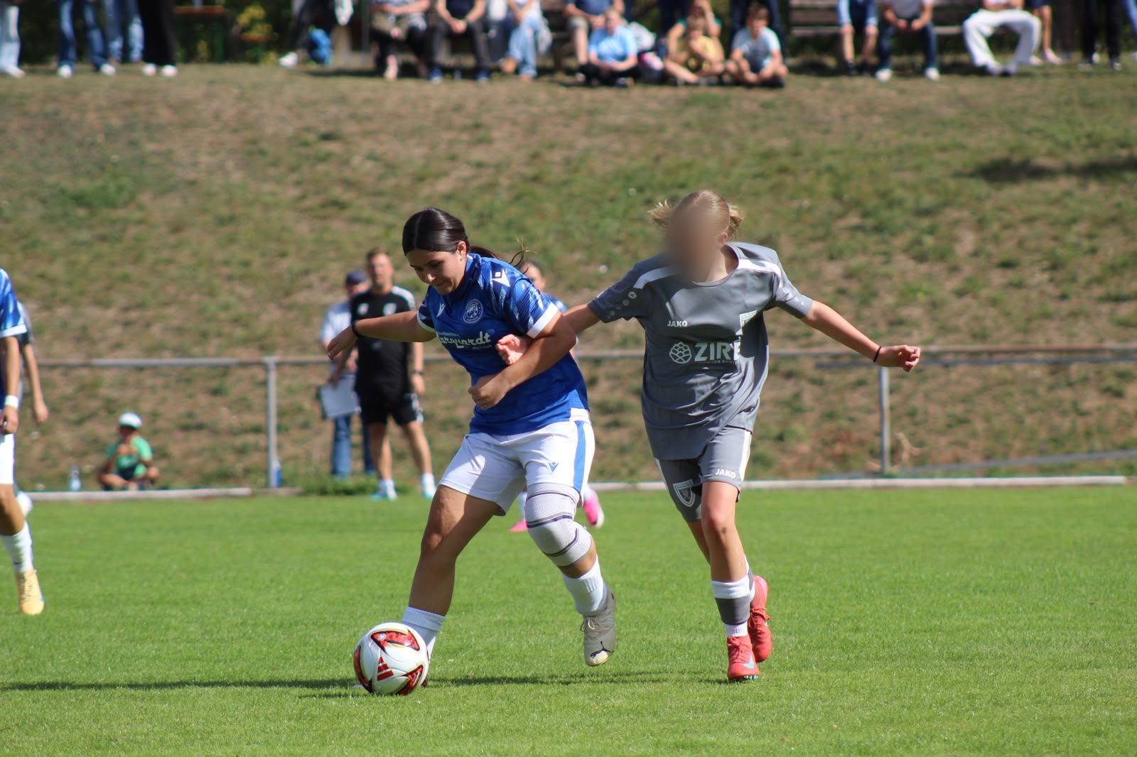 Spielerin des FC Panthers im blauen Trikot schirmt den Ball gegen eine Gegenspielerin in Grau ab, im Hintergrund Zuschauerinnen und Zuschauer am Hang.