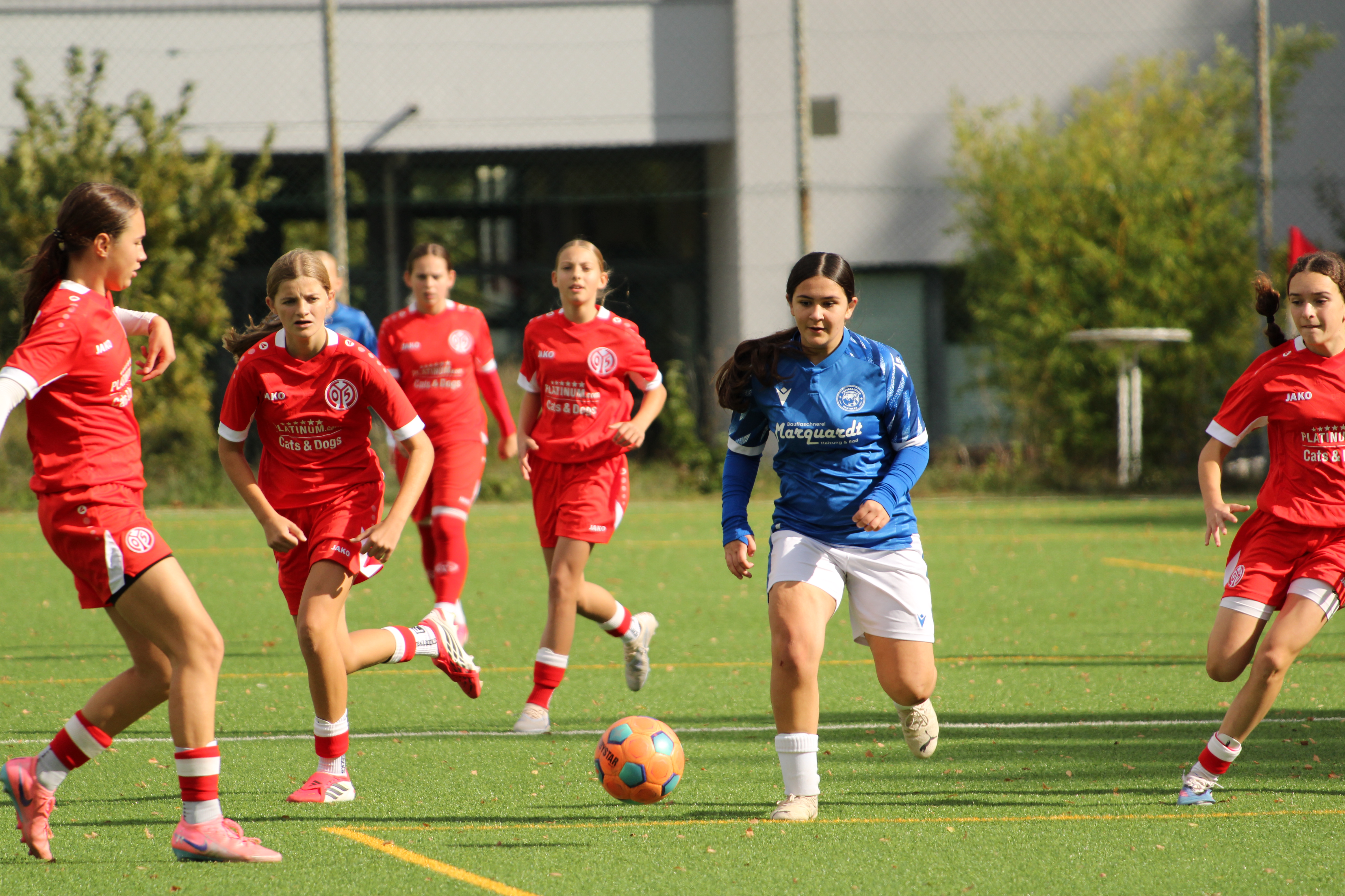 Spielerin in blauem Trikot läuft dem Ball nach, umgeben von mehreren Spielerinnen in roten Trikots.