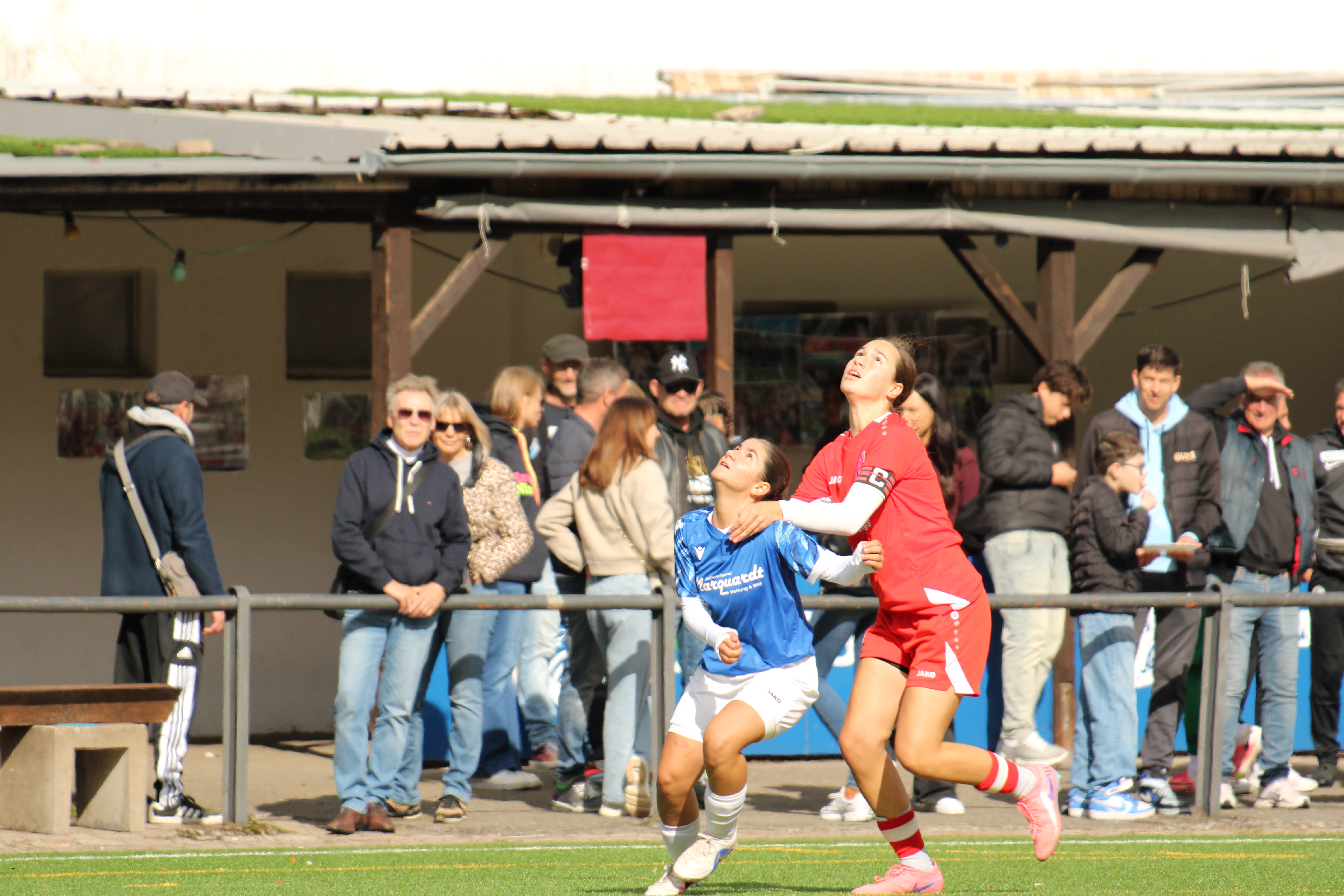 Eine Spielerin in Blau und eine Spielerin in Rot schauen im Zweikampf nach oben zum Ball, dahinter Zuschauende am Geländer.