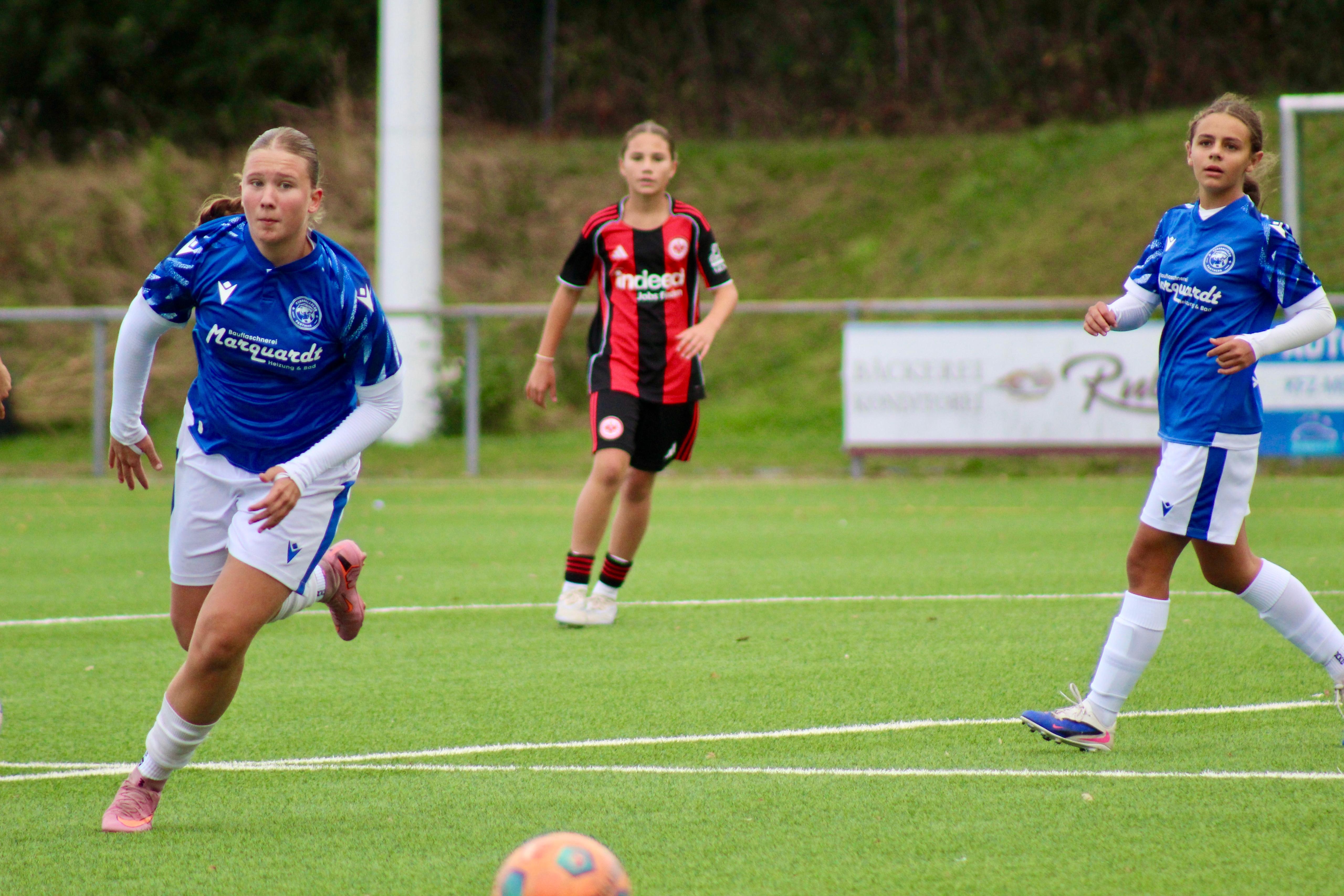 Zwei Spielerinnen der Panthers in blauen Trikots laufen dem Ball nach, im Hintergrund eine Spielerin von Eintracht Frankfurt.