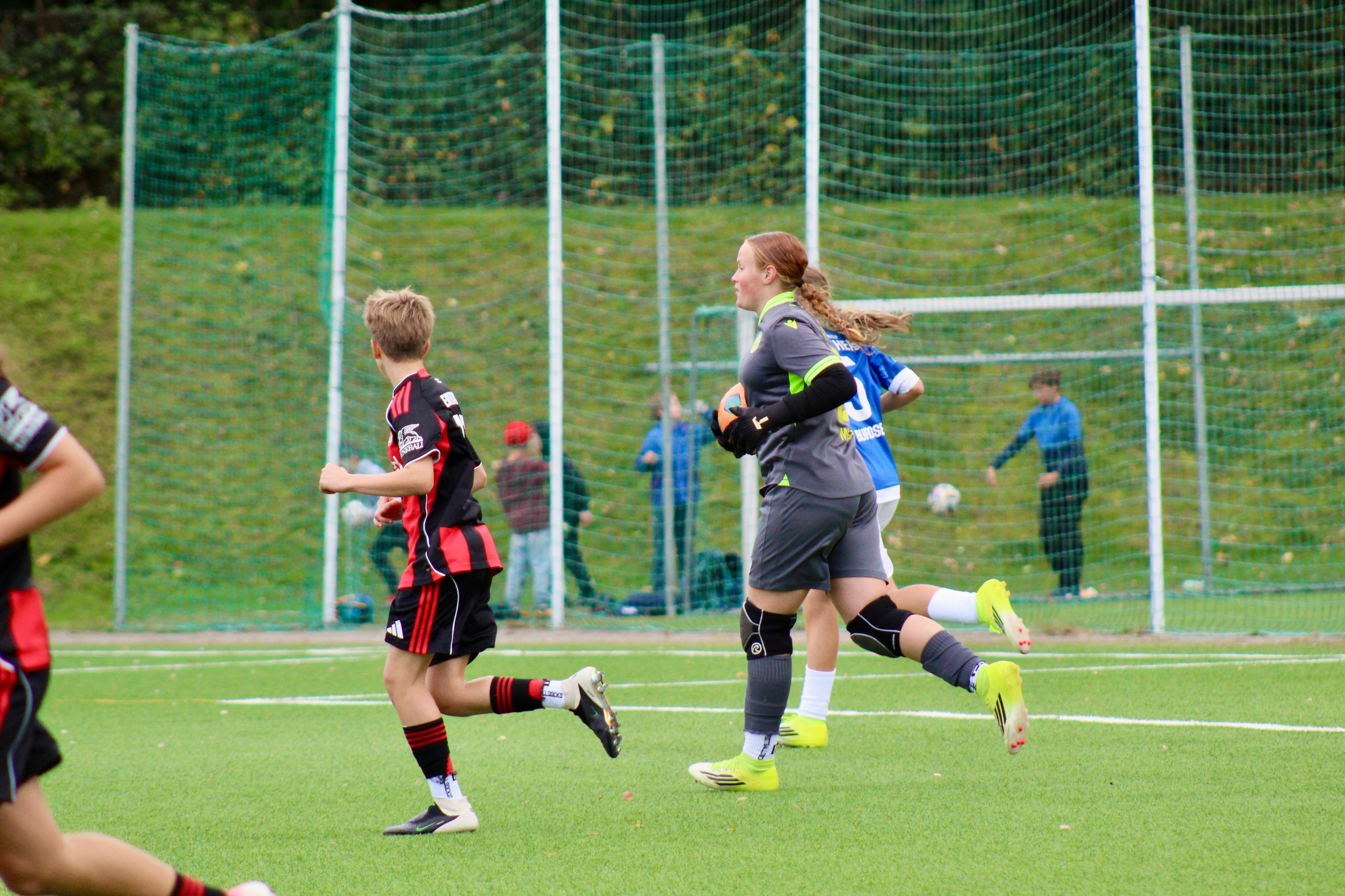 Die Torhüterin der Panthers in grauem Trikot läuft mit dem Ball in den Händen über den Kunstrasen, daneben Spielerinnen beider Teams.