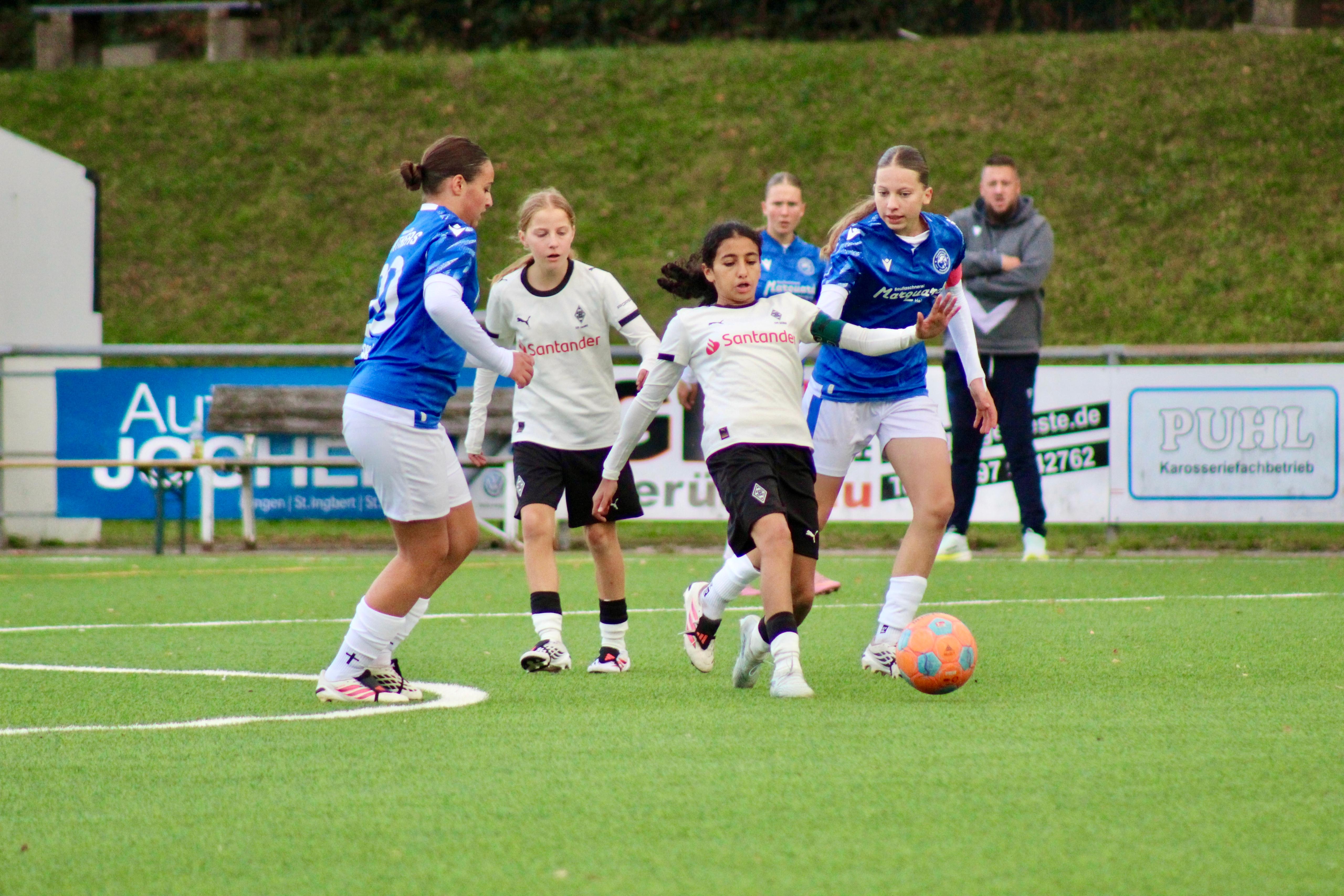 Spielerin in weißem Trikot am Ball, umringt von zwei Spielerinnen in blauen Trikots, eine davon mit Kapitänsbinde
