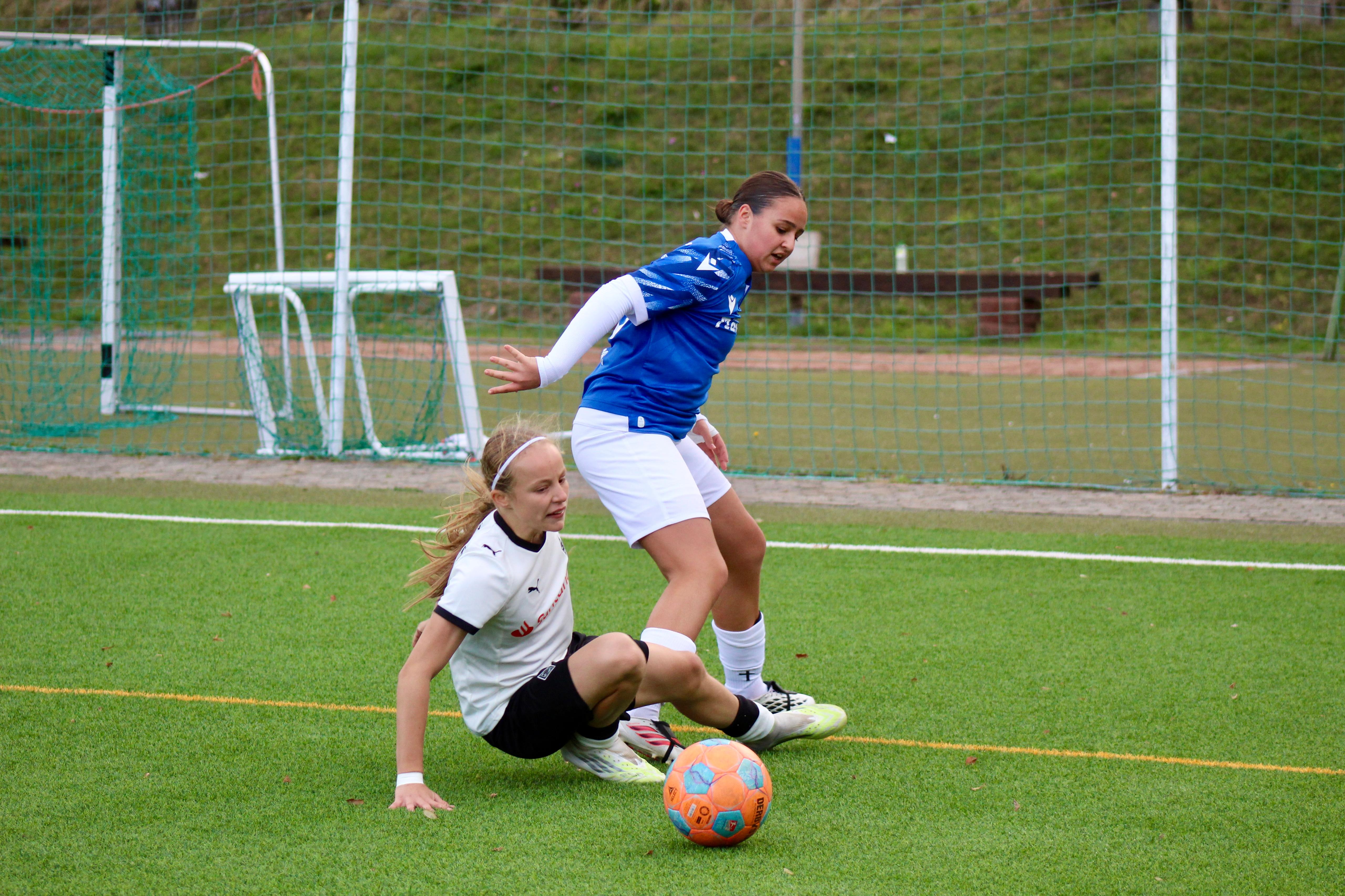 Spielerin in blauem Trikot behauptet den Ball an der Seitenlinie, während eine Spielerin in weißem Trikot zur Grätsche ansetzt