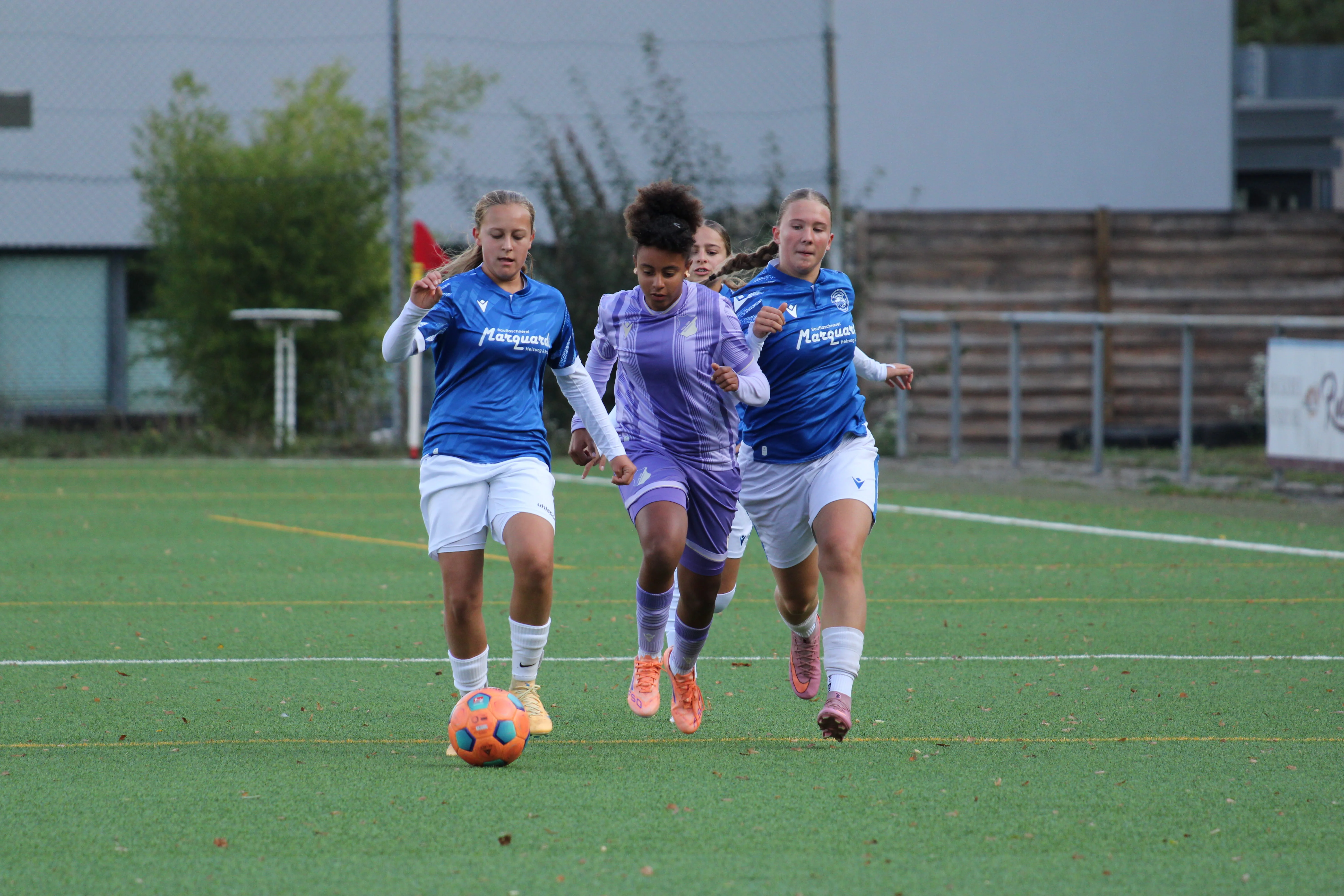 Spielerin in lila Trikot läuft zwischen zwei Spielerinnen in blauen Trikots dem Ball hinterher