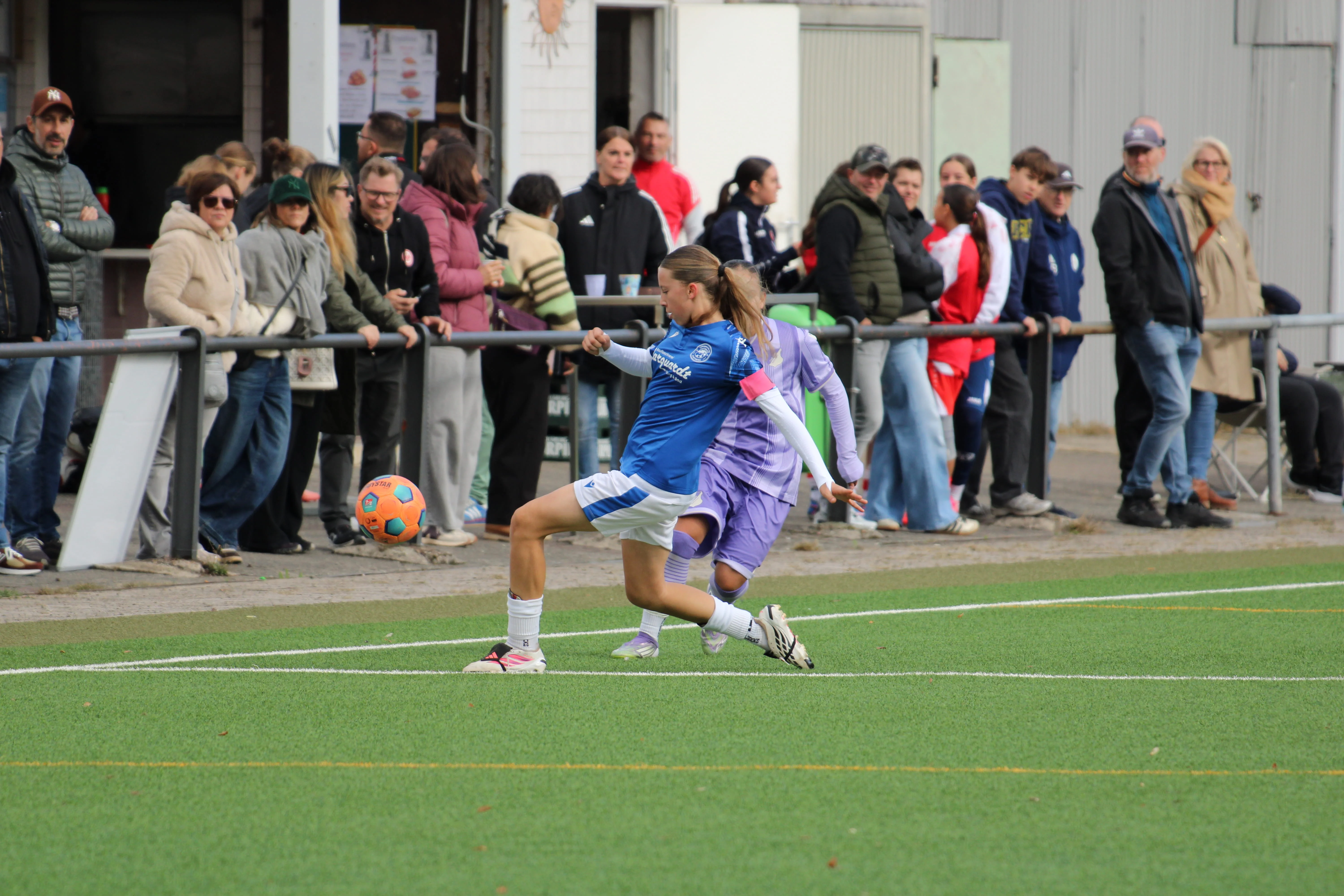 Spielerin im blauen Trikot mit Kapitänsbinde spielt den Ball an der Seitenlinie, dahinter eine Gegenspielerin in Lila und Zuschauer am Geländer